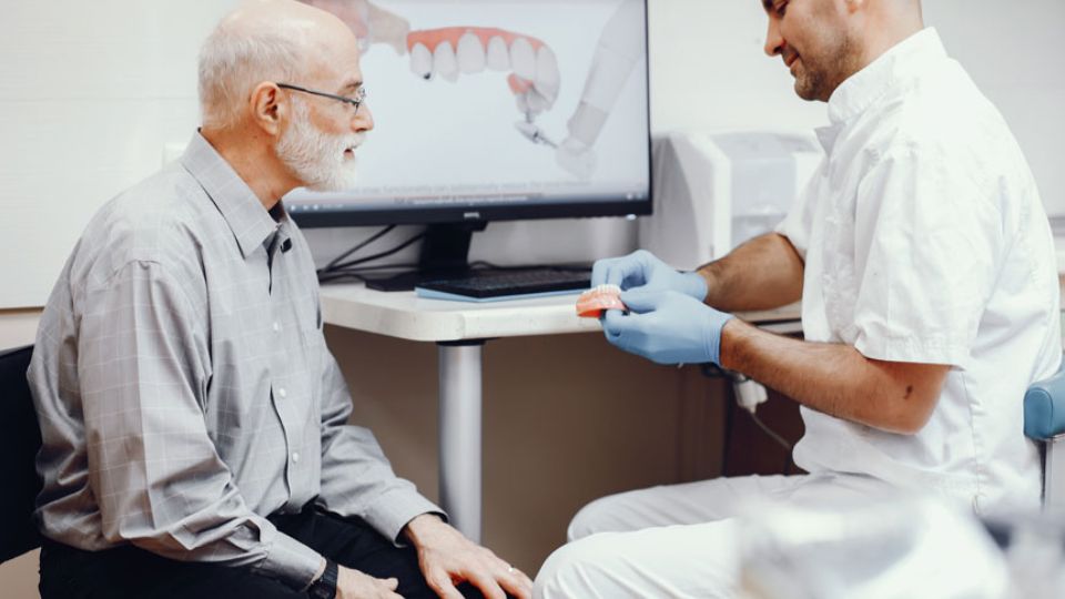 Senior Man Consulting With A Dentist Who Is Holding Dental Prosthetics